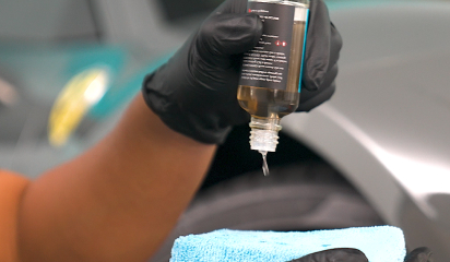 A gloved hand pours a clear liquid from a small bottle onto a blue microfiber applicator pad. The applicator is held near a car wheel area, suggesting preparation for applying a detailing or protective coating.