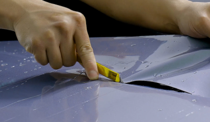 Hands using a precision blade to trim a sheet of glossy colored paint protection film on a car hood.
