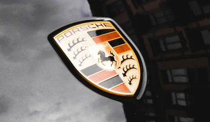 Close-up of a Porsche emblem on a sleek black car hood, reflecting a cloudy sky and nearby buildings.