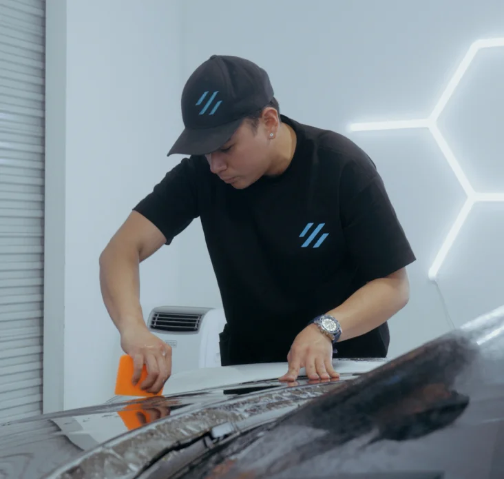Automotive technician applying protective film to a vehicle hood with a squeegee inside a detailing studio.