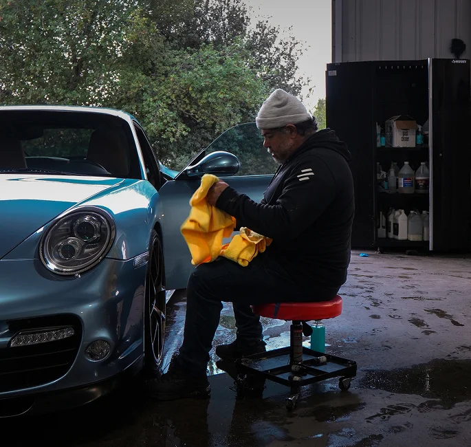 Detailing technician preparing a yellow microfiber towel next to a glossy blue Porsche undergoing protective coating.